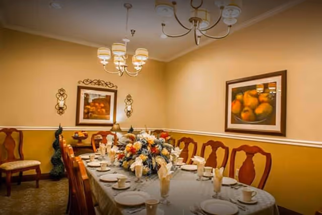 A formal dining room with a long table set for a meal, featuring white plates, cups, and napkins. The table has a large floral centerpiece. The room has yellow and beige walls with two framed paintings of fruit and decorative wall sconces. Wooden chairs with cushioned seats surround the table, and a chandelier with multiple lights hangs from the ceiling.