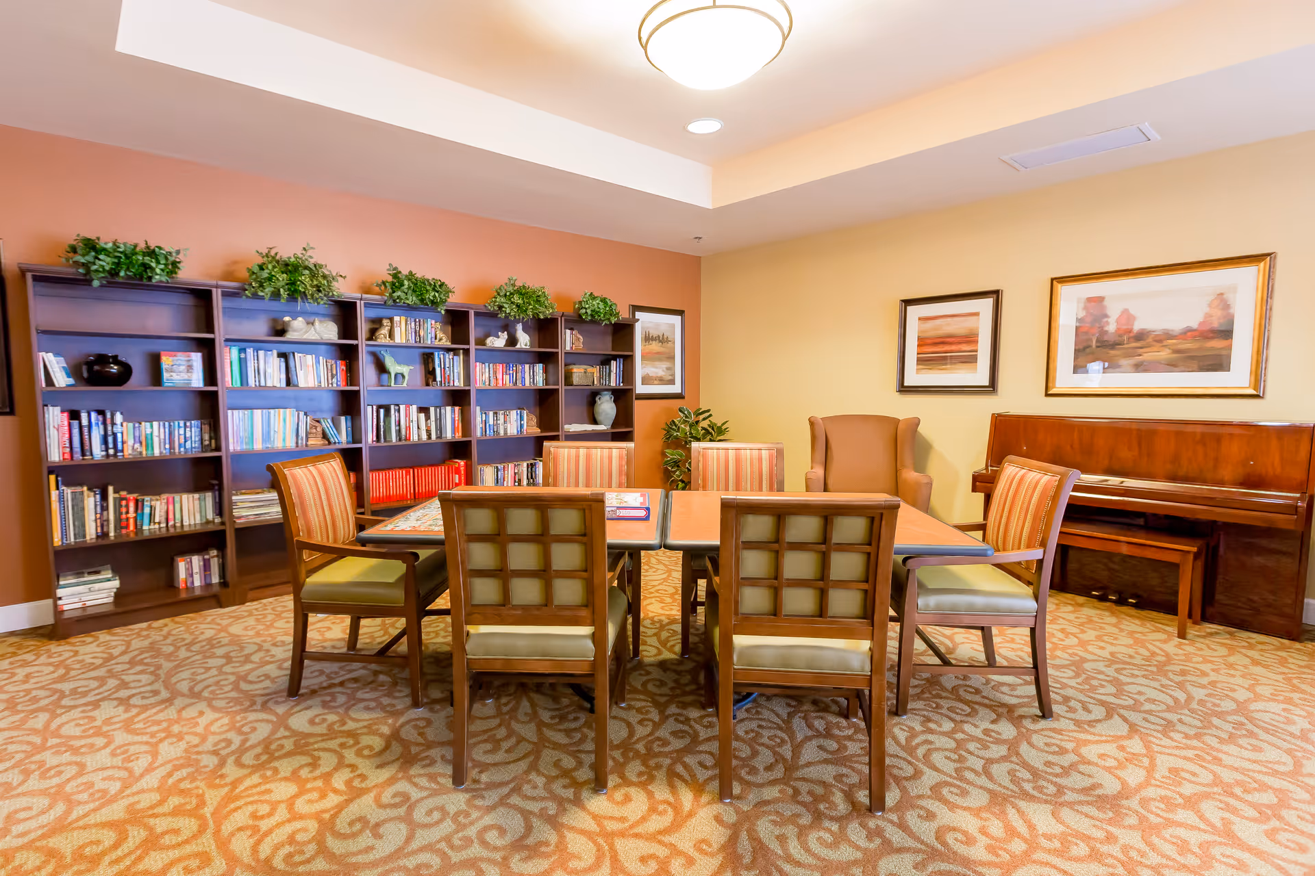 A community sitting room with a central table and chairs, bookshelves along the back wall, and a piano.