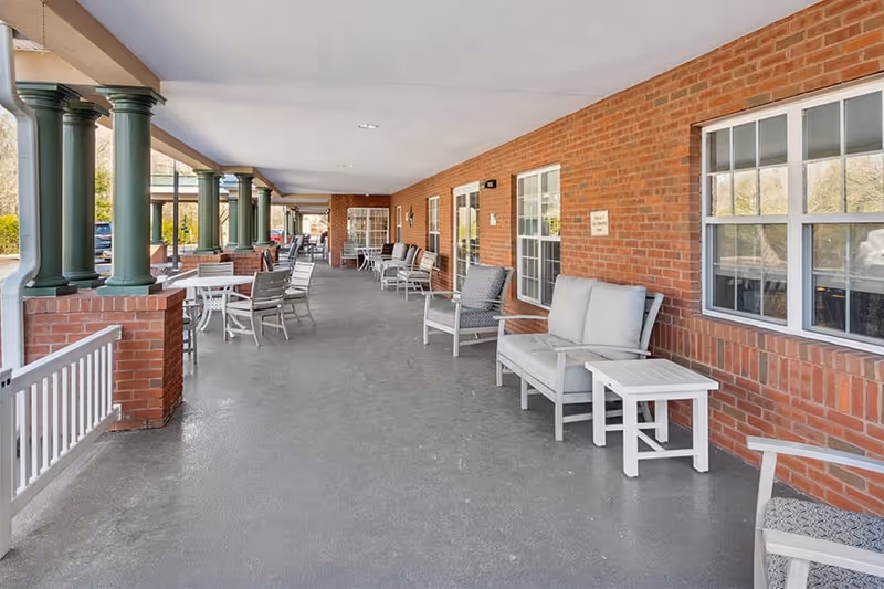 A brick porch with multiple seating areas, featuring white chairs and tables, and green columns supporting the roof.