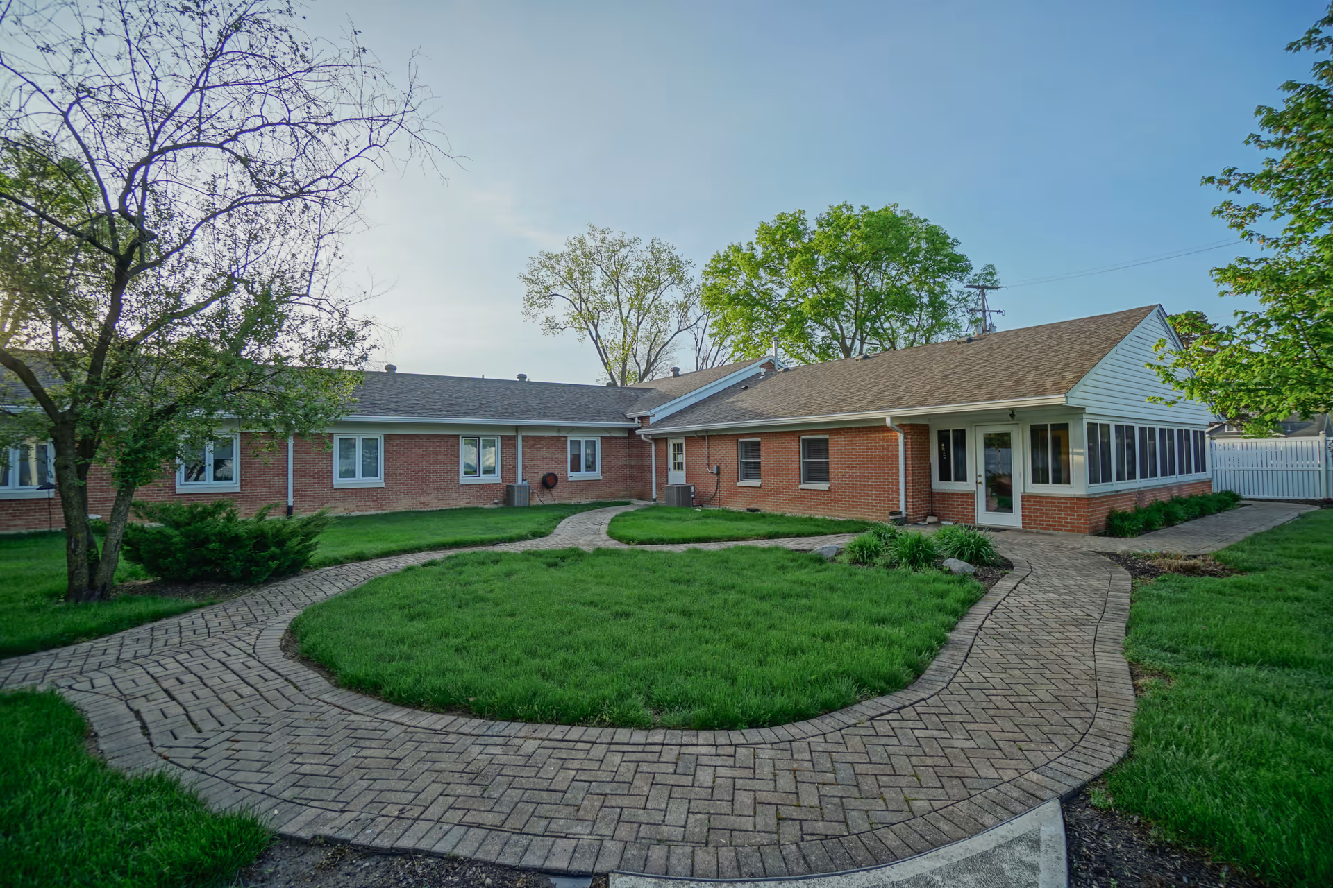 A single-story brick building with multiple windows and a shingled roof, surrounded by a green lawn and trees. A paved walkway curves through the grass leading to the building's entrances under a clear sky.