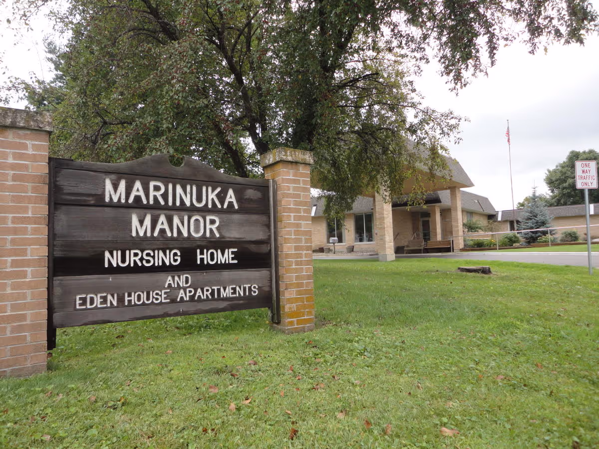 A wooden sign reading "Marinuka Manor Nursing Home and Eden House Apartments" on a lawn in front of the facility building and entrance.