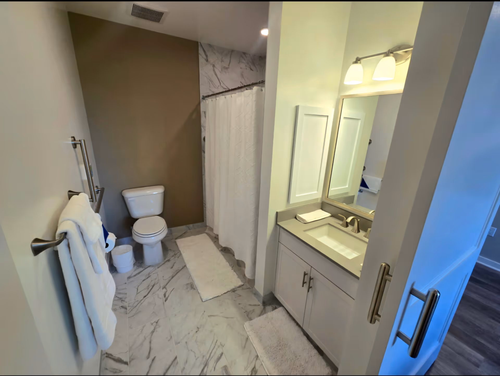 A modern bathroom with a white toilet, a shower with a white curtain, a vanity with a sink, a large mirror, and two light fixtures above the mirror. The floor is covered with white and gray marble tiles, and there are white bath mats on the floor. White towels hang on a towel rack on the left wall.