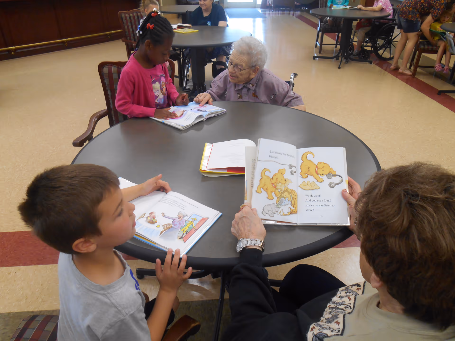 An elderly woman in a wheelchair and a young girl sit at a round table reading books together. Nearby, a young boy and an adult woman are also seated at the table, each holding a book. Other people are visible in the background sitting at tables in a spacious room with a tiled floor.