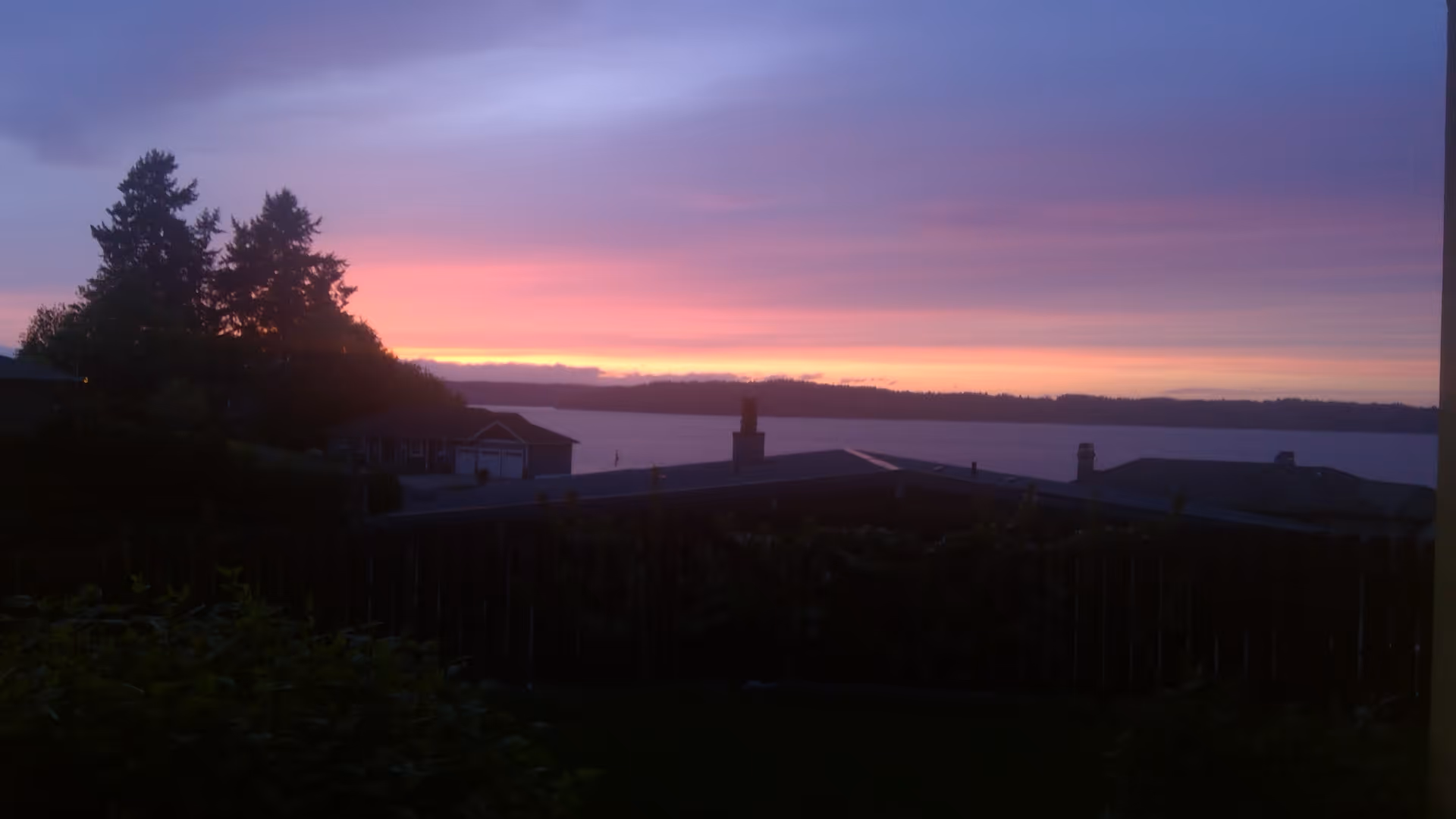 View of a sunset over a body of water with silhouetted trees, rooftops, and a wooden fence in the foreground.