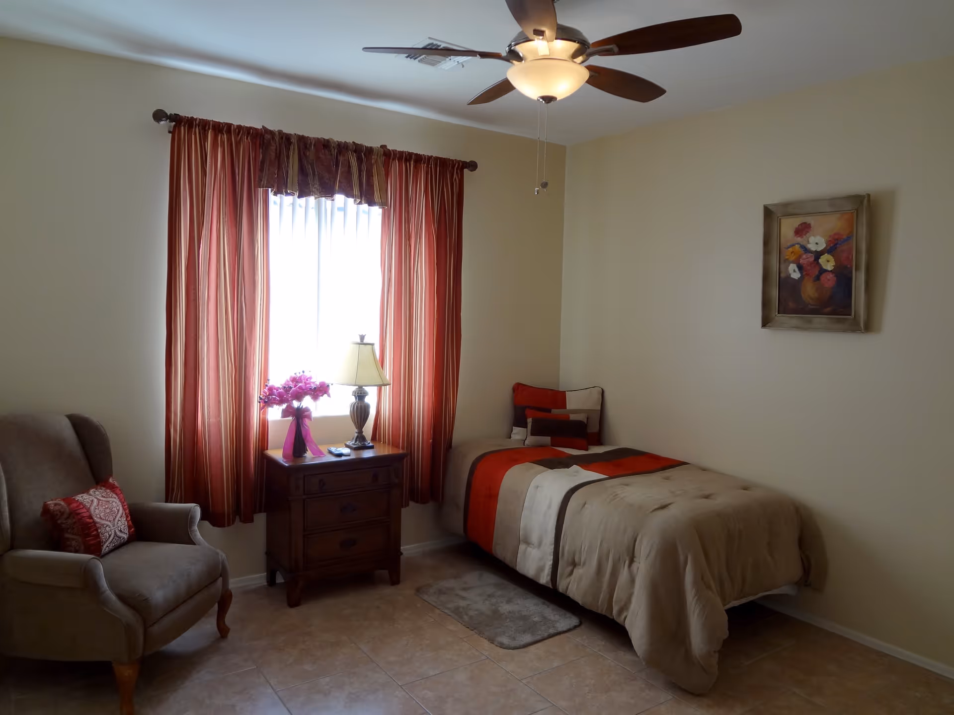 A small bedroom with a single bed covered in a beige, red, and brown striped comforter. Next to the bed is a wooden nightstand with a lamp and a vase of pink flowers. A window with red and beige striped curtains is behind the nightstand. To the left is a beige upholstered armchair with a red decorative pillow. A ceiling fan with a light is mounted on the ceiling, and a framed painting of flowers hangs on the wall above the bed.