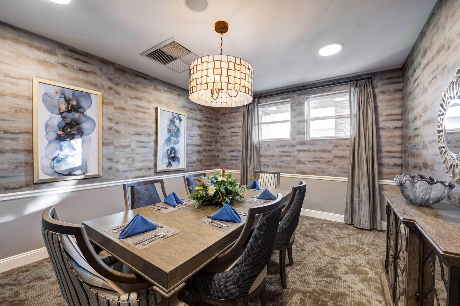A well-decorated dining room with a rectangular wooden table set for six people. The table has blue folded napkins, silverware, and a floral centerpiece. The room features patterned wallpaper, two framed abstract paintings, a large decorative mirror, and a sideboard with a decorative bowl. There are two windows with curtains and a ceiling light fixture above the table.