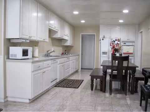 A clean kitchen with white cabinets and countertops, a microwave on the left counter, a sink, and a stove. There is a dark wooden dining table with matching chairs and a bench on the right side. The floor is tiled, and the ceiling has recessed lighting. A refrigerator with magnets and a door are visible at the back.