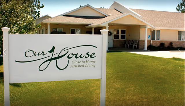 White sign reading 'Our House Close to Home Assisted Living' on a grassy lawn in front of a single-story assisted living building.