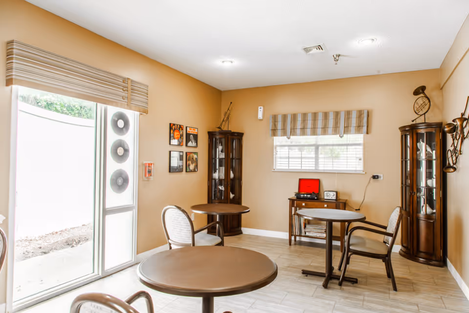 Bright common room with round tables and chairs, two glass display cabinets, a small radio console and a window.