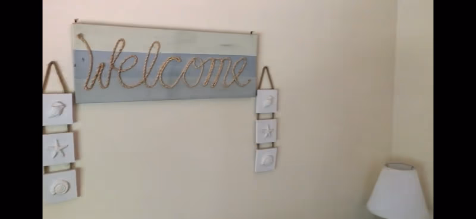 Pale interior wall with a decorative 'Welcome' sign and hanging sea-themed tiles, and a lamp visible at the right edge.