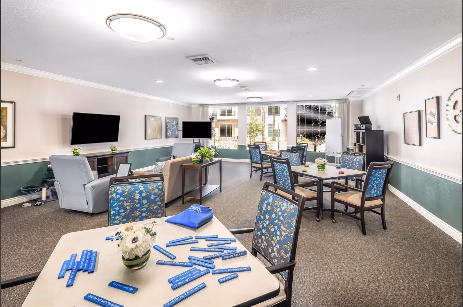 Bright communal activity room with tables and floral-upholstered chairs, sofas, TVs, and large windows.