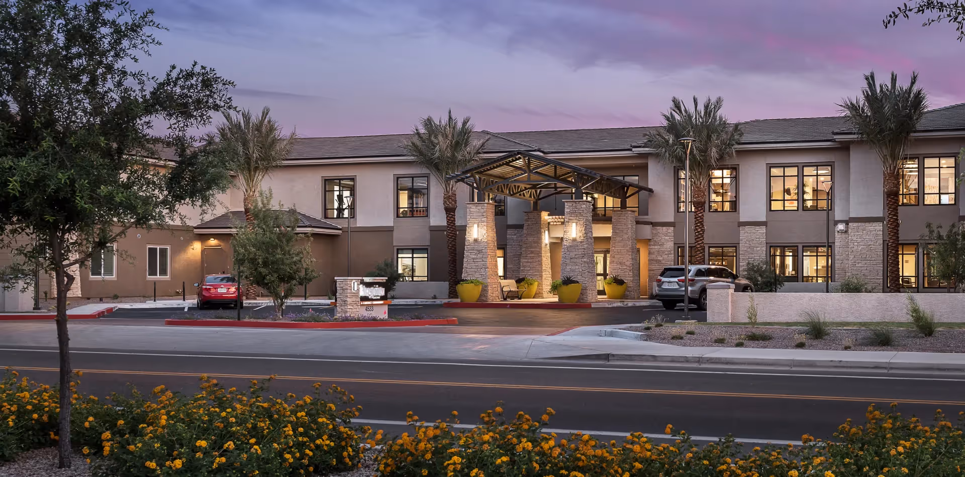 Front exterior of a two-story senior living building at dusk with a covered entrance, palm trees, parked cars, and landscaped foreground.
