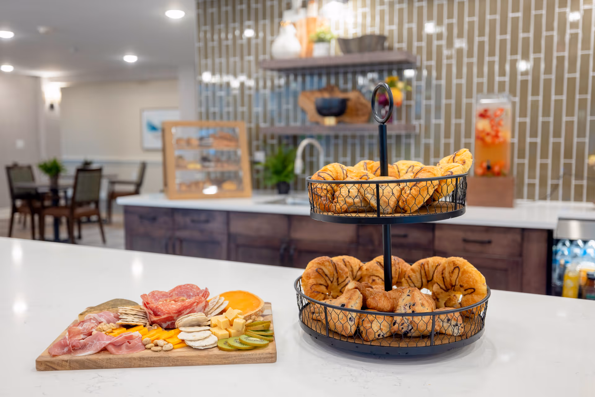 A kitchen counter with a two-tiered basket filled with assorted pastries and a wooden board with an assortment of sliced meats, cheeses, crackers, and kiwi slices. In the background, there is a kitchen area with a sink, shelves with decorative items, and a dining area with tables and chairs.