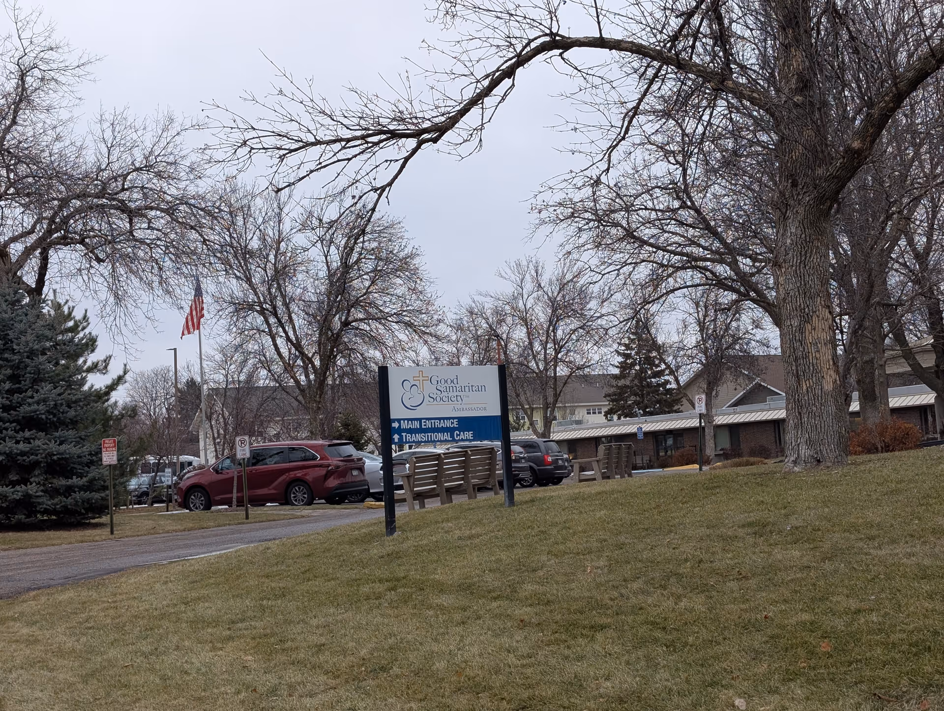 Outdoor view of Good Samaritan Ambassador facility with a sign indicating main entrance and transitional care. Several cars are parked near the building, leafless trees and an American flag are visible under a cloudy sky.