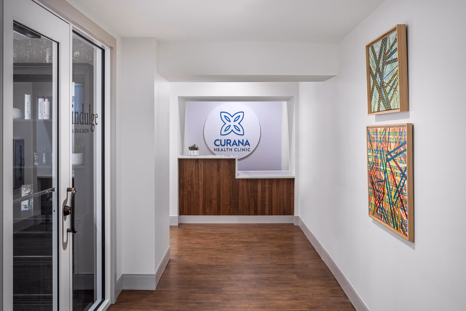 A clean hallway leading to a Curana Health Clinic reception desk with framed artwork on the right wall and glass doors on the left.