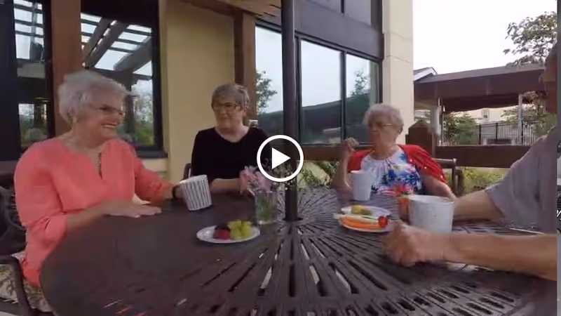 Four elderly people sitting around a round outdoor table with drinks and plates of fruit, engaged in conversation outside a building with large windows.
