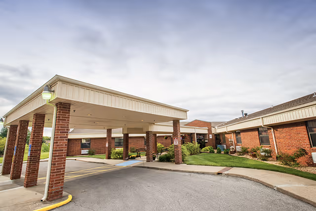 Exterior view of Pleasant Hill Health and Rehabilitation Center showing a covered entrance with brick pillars, a driveway, and a well-maintained lawn with shrubs and bushes under a cloudy sky.