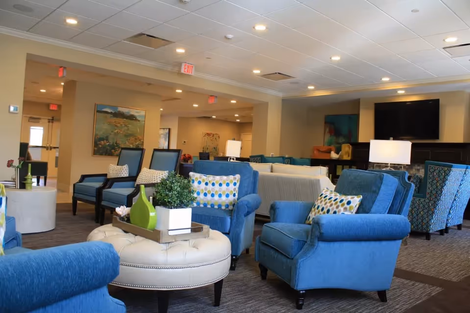 Bright communal lounge with blue armchairs, a round ottoman, decorative pillows and a wall-mounted TV.