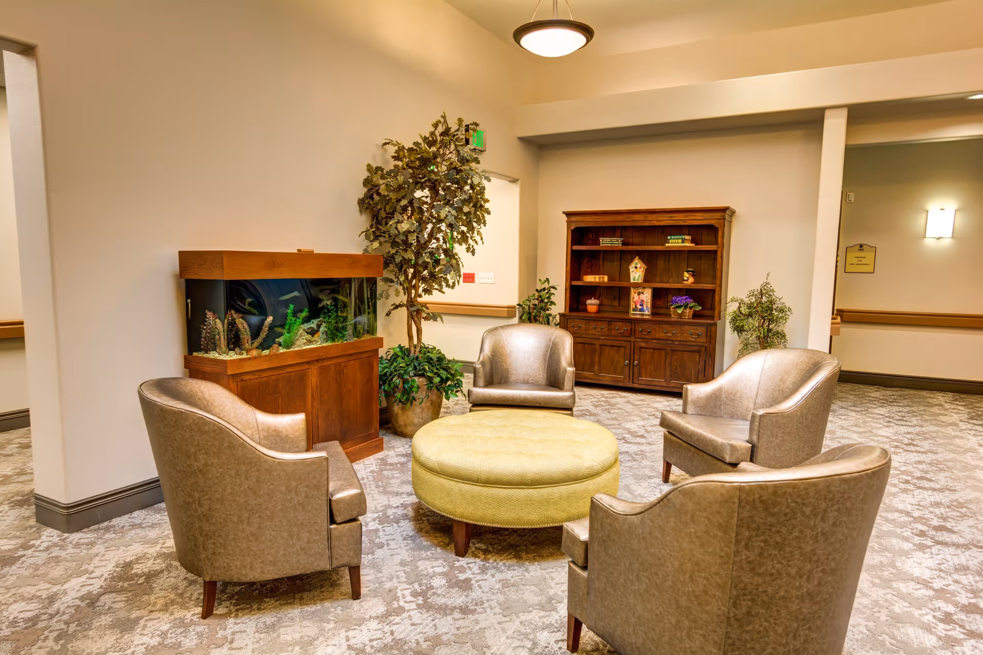 A cozy seating area with four beige armchairs arranged around a round green ottoman. Behind the chairs is a wooden cabinet with shelves holding decorative items and books. To the left, there is a large fish tank on a wooden stand and a tall potted plant. The room has soft lighting and neutral-colored walls and carpet.