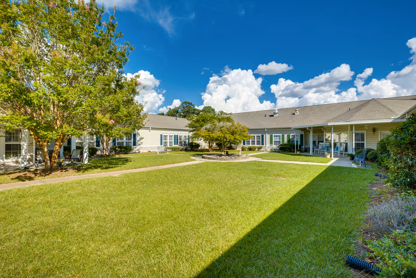 A sunny outdoor courtyard area at The Legacy of Lexington featuring a well-maintained green lawn, a central tree surrounded by a small stone border, and a building with a covered porch and rocking chairs. The sky is bright blue with scattered white clouds.