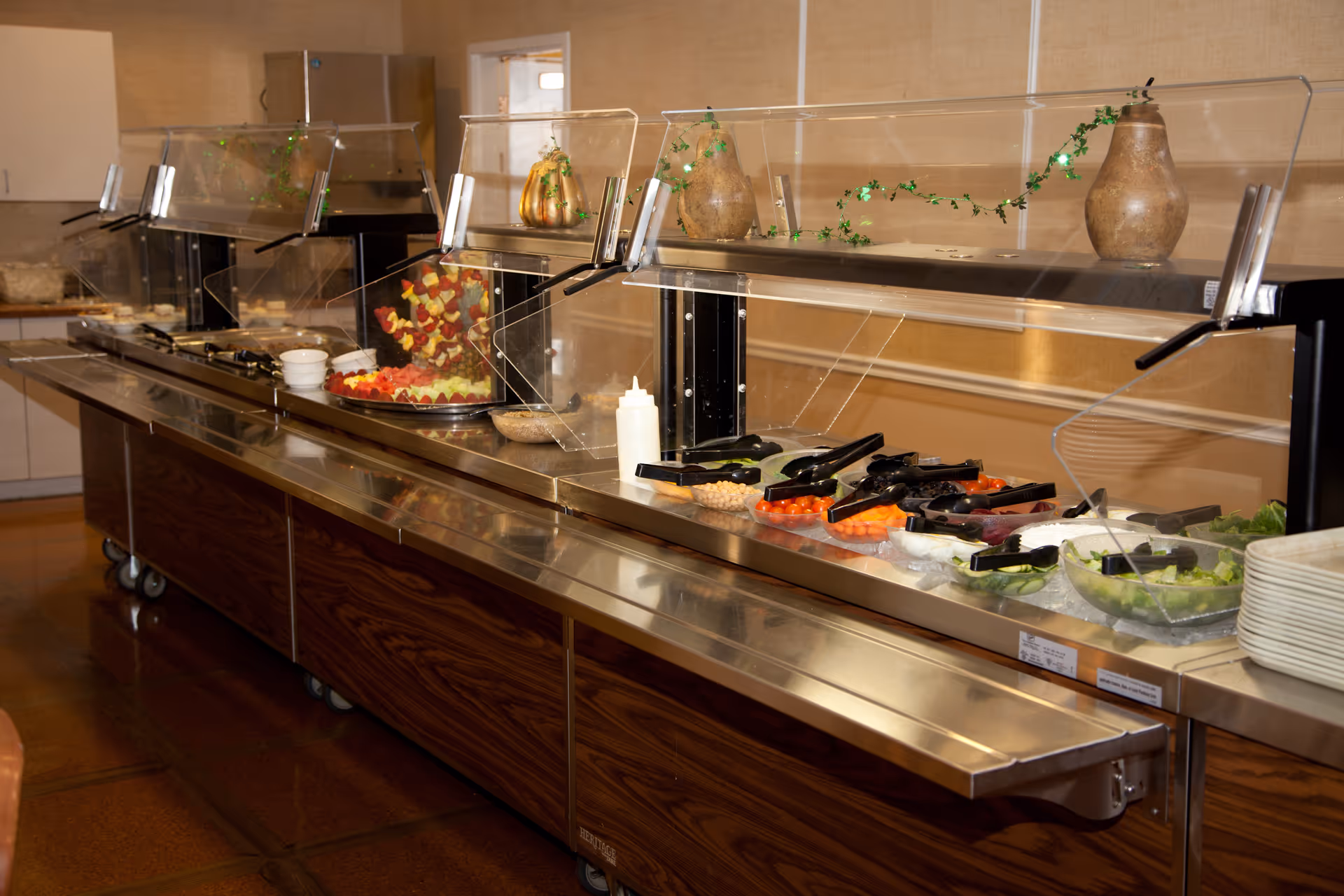 A buffet-style salad bar with various fresh vegetables and fruits displayed in bowls, protected by a clear sneeze guard. The counter has a stainless steel surface with wooden paneling underneath, and decorative vases with greenery are placed on top of the sneeze guard.
