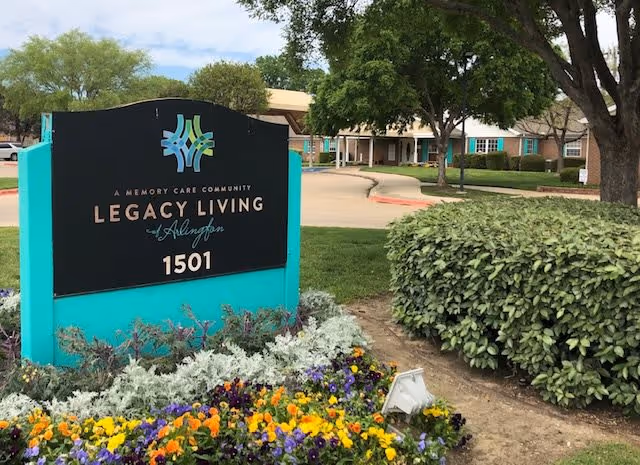 Outdoor view of the entrance area of Legacy Living Memory Care community, featuring a large sign with the facility name and address 1501, surrounded by colorful flowers, bushes, trees, and a driveway leading to the building in the background.