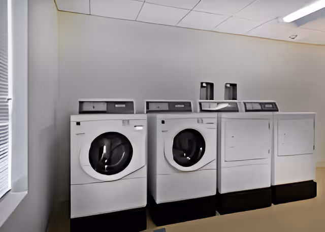 A laundry room with two front-loading washing machines and two top-loading dryers lined up against a plain white wall. There is a window with blinds partially visible on the left side, and a fluorescent light fixture on the ceiling.