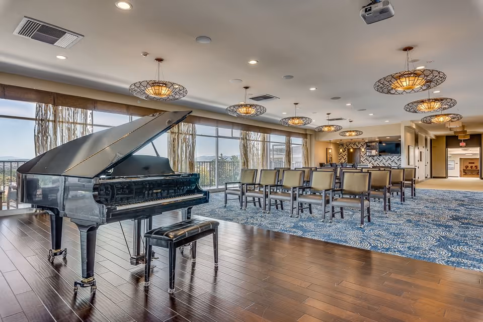 Sunlit communal activity room with a grand piano, rows of chairs, and decorative hanging lights overlooking large windows.