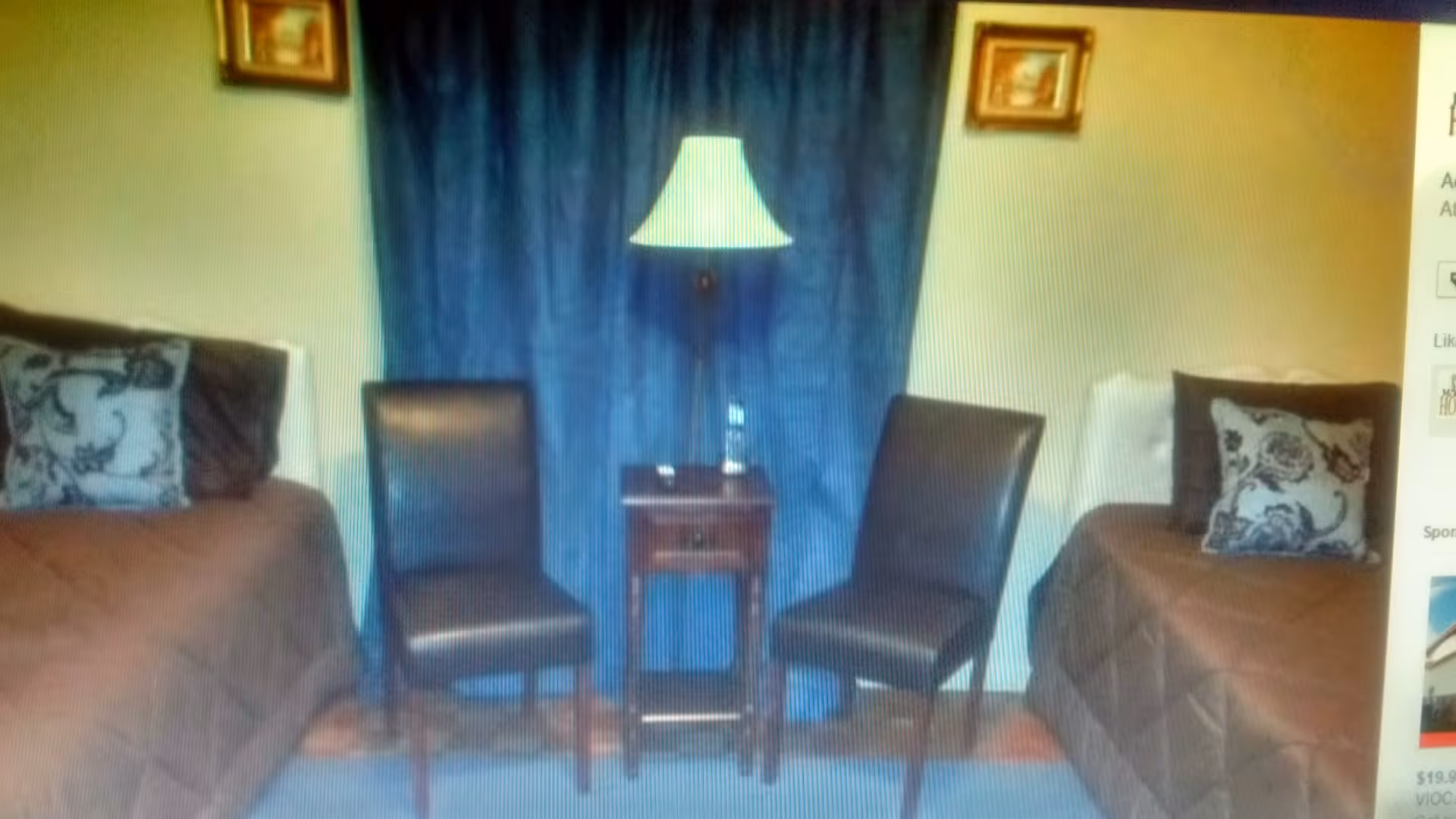 A cozy room with two single beds on either side, each with brown bedding and decorative pillows. Between the beds are two dark brown chairs and a small wooden table with a lamp on it. The background features a blue curtain and two framed pictures on the wall.
