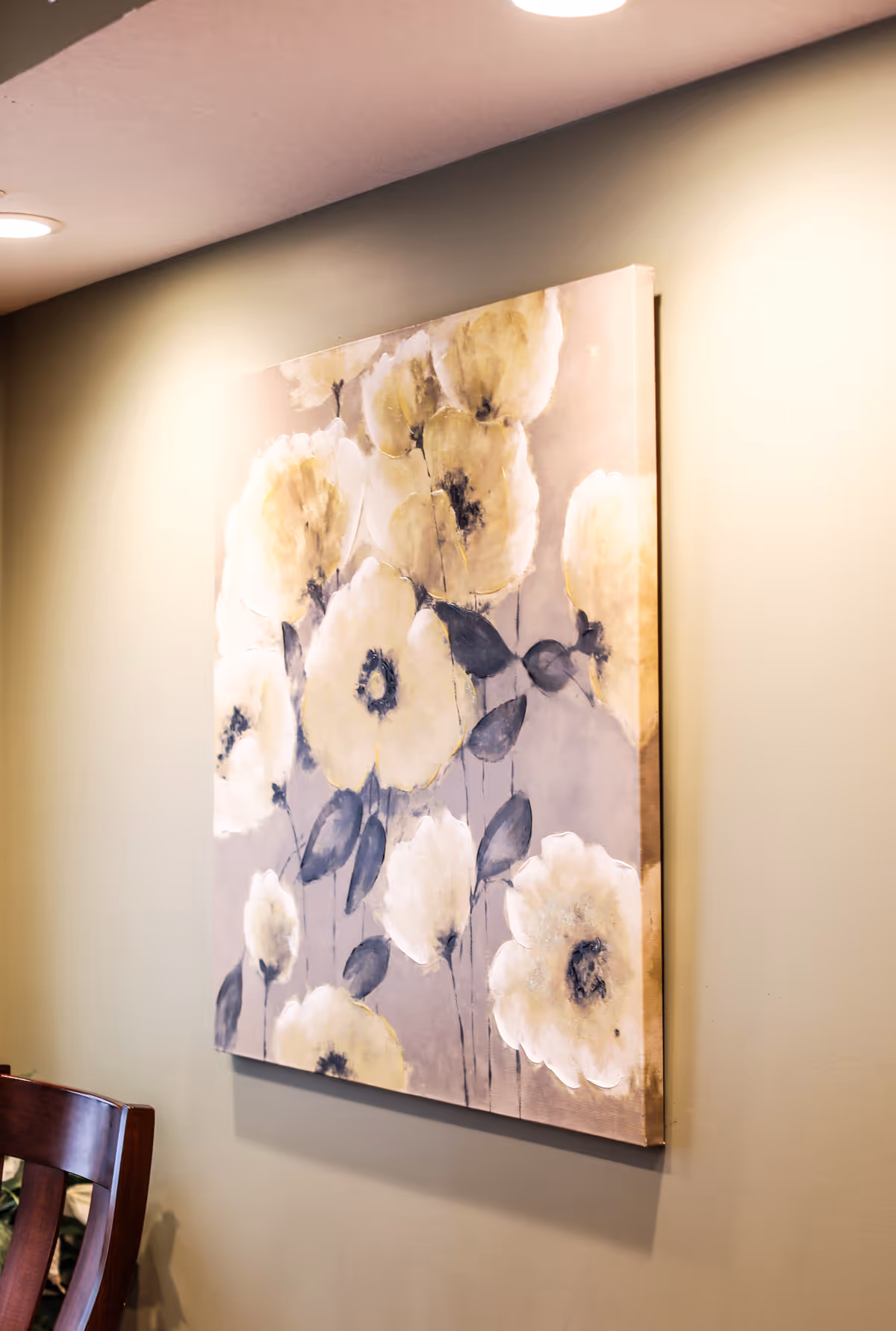 A close-up view of a wall-mounted painting featuring large white and yellow flowers with dark centers and leaves on a muted background, illuminated by ceiling lights. Part of a wooden chair is visible at the bottom left corner.