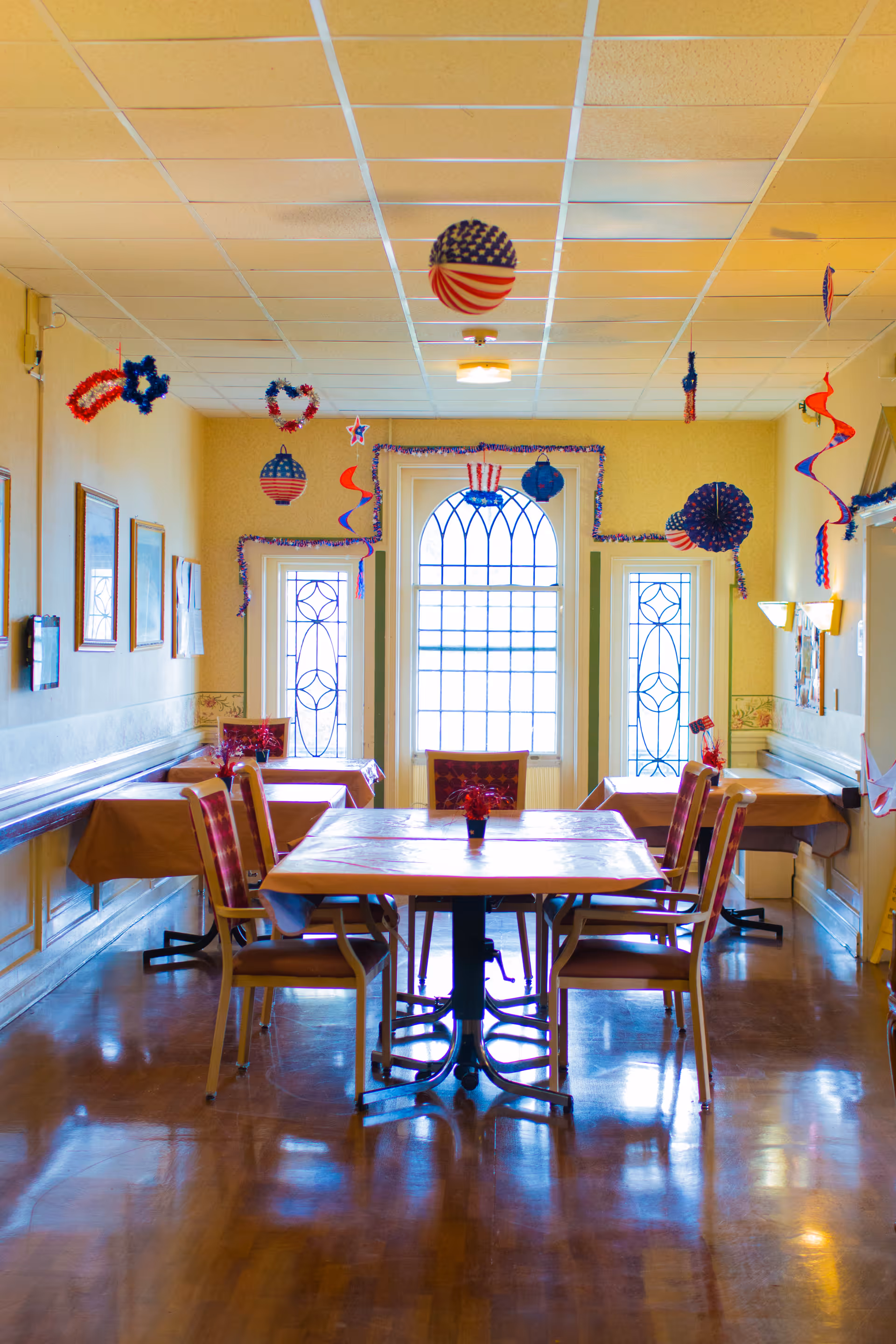 A dining room with tables and chairs arranged neatly. The room is decorated with patriotic-themed hanging decorations including stars, hearts, and circular ornaments in red, white, and blue. There are three tall windows with decorative glass panes letting in natural light. The floor is polished wood, and the walls have framed pictures and wall sconces.