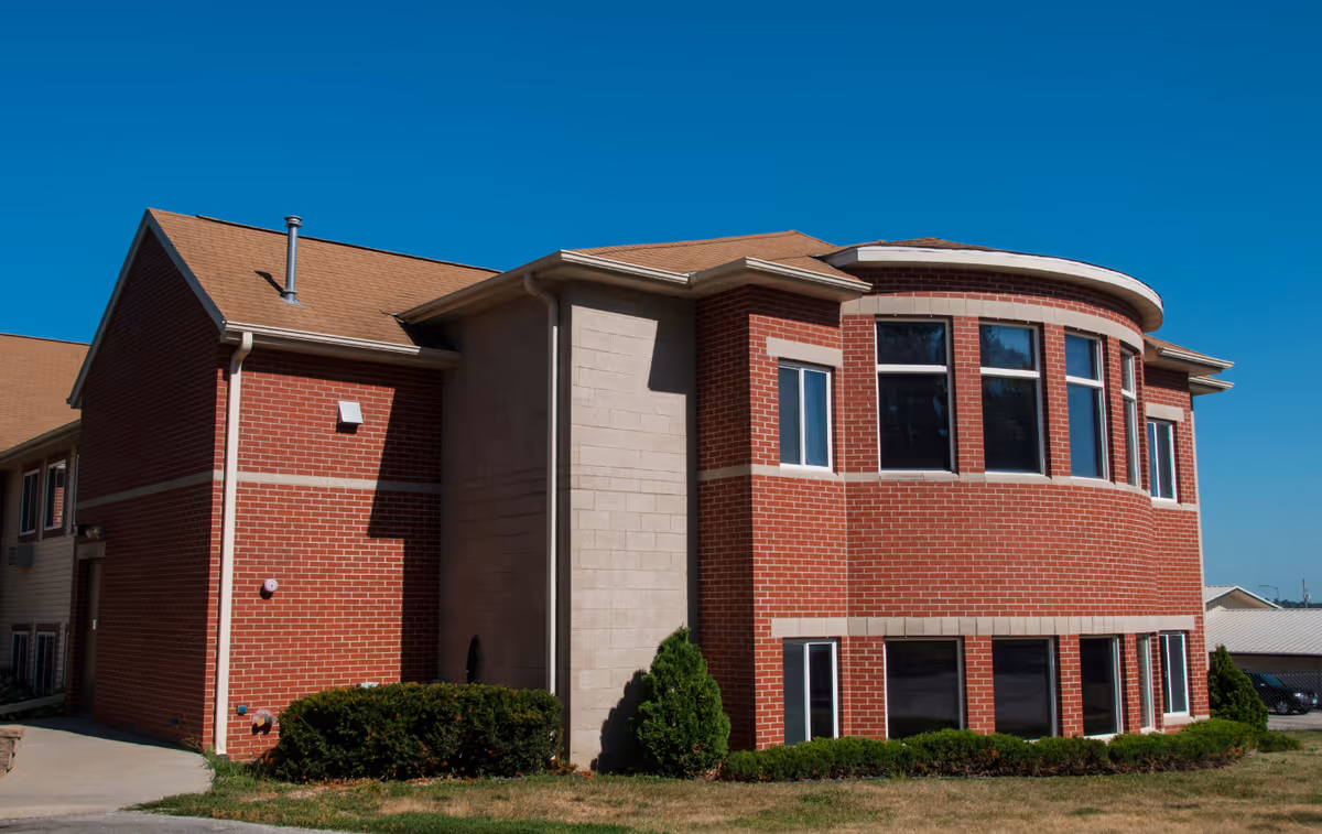 Exterior view of a two-story brick building with a rounded front section and multiple windows under a clear blue sky. The building has a tan roof and is surrounded by small bushes and grass.
