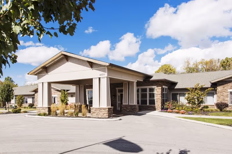 Front entrance of a single-story assisted living building with a covered porte-cochere, stone accents and landscaped grounds under a blue sky.