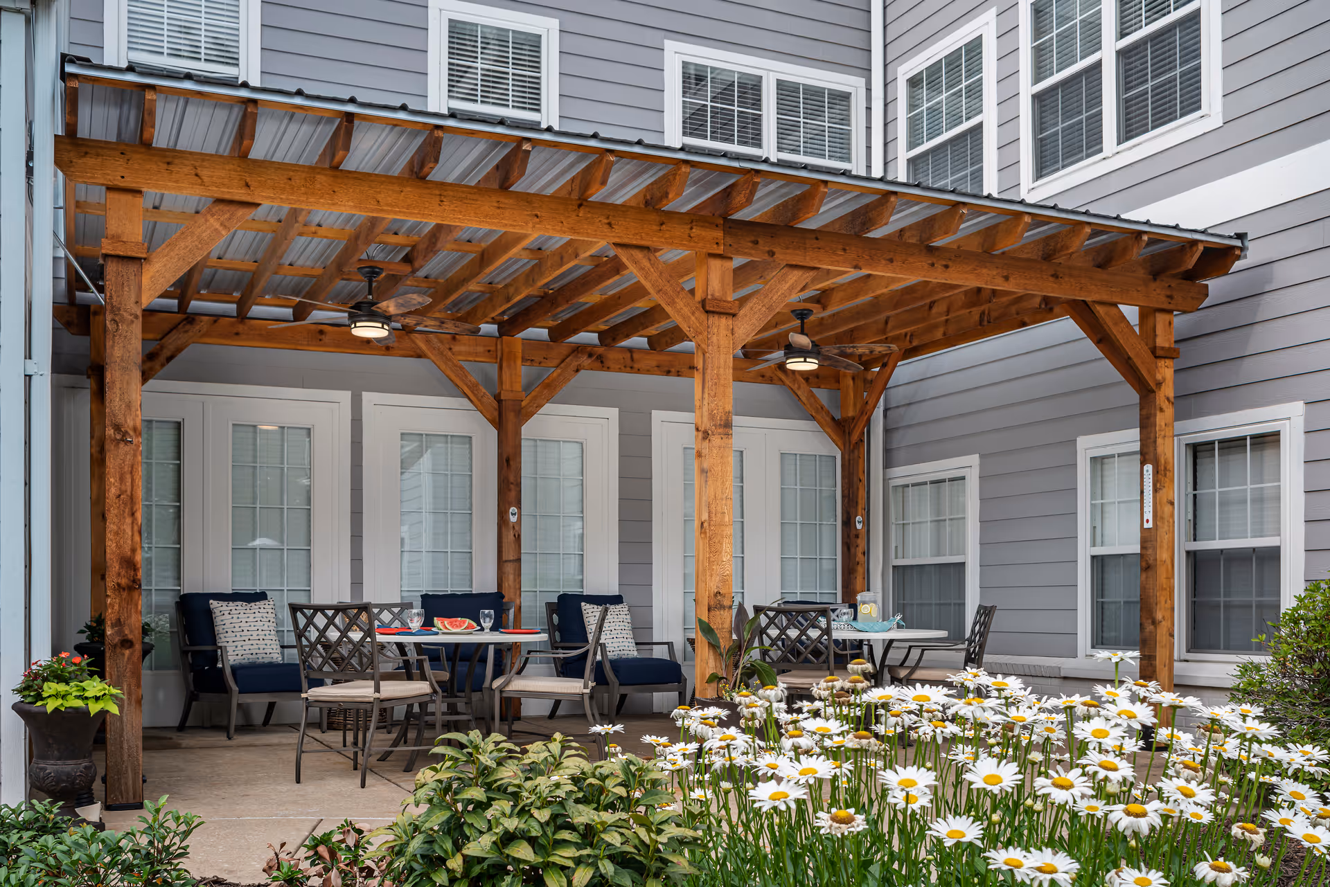 Outdoor patio area at The Grove at Midtown with a wooden pergola covering several tables and chairs. The patio is adjacent to a gray building with white-trimmed windows and doors. There are potted plants and a flower bed with white daisies in the foreground.
