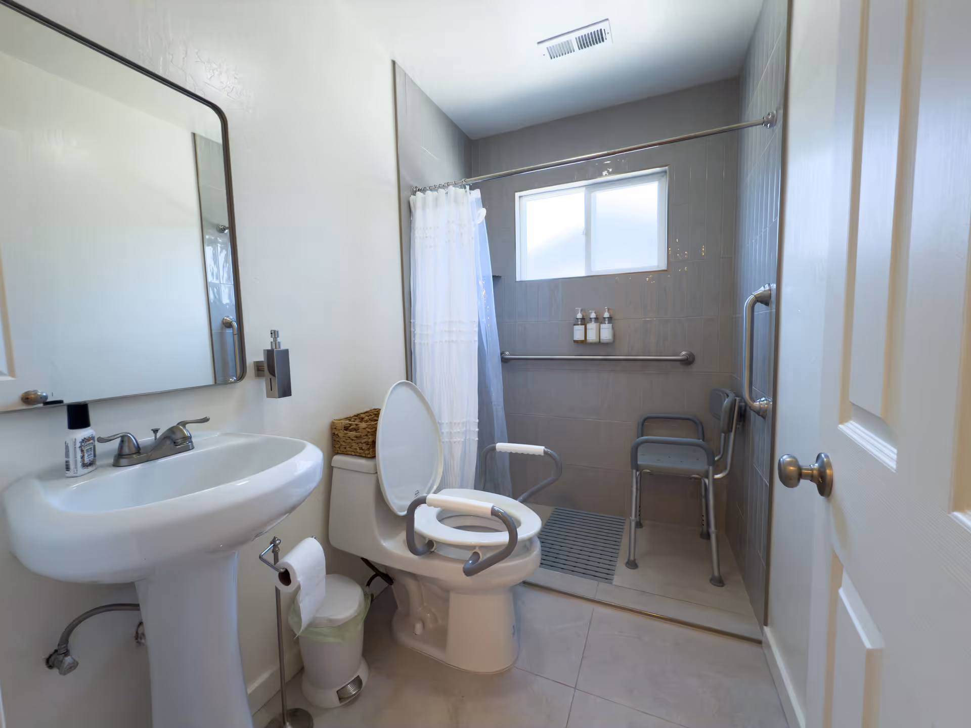 A clean and accessible bathroom with a pedestal sink, a toilet equipped with safety rails, and a walk-in shower area with a shower chair, grab bars, and a window. The shower has a white curtain and mounted soap dispensers.