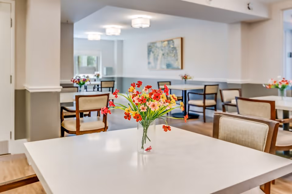 A bright and clean dining room in a senior living facility with multiple tables and chairs. Each table has a vase with colorful flowers, and there is a painting on the wall. The room has soft lighting and a welcoming atmosphere.