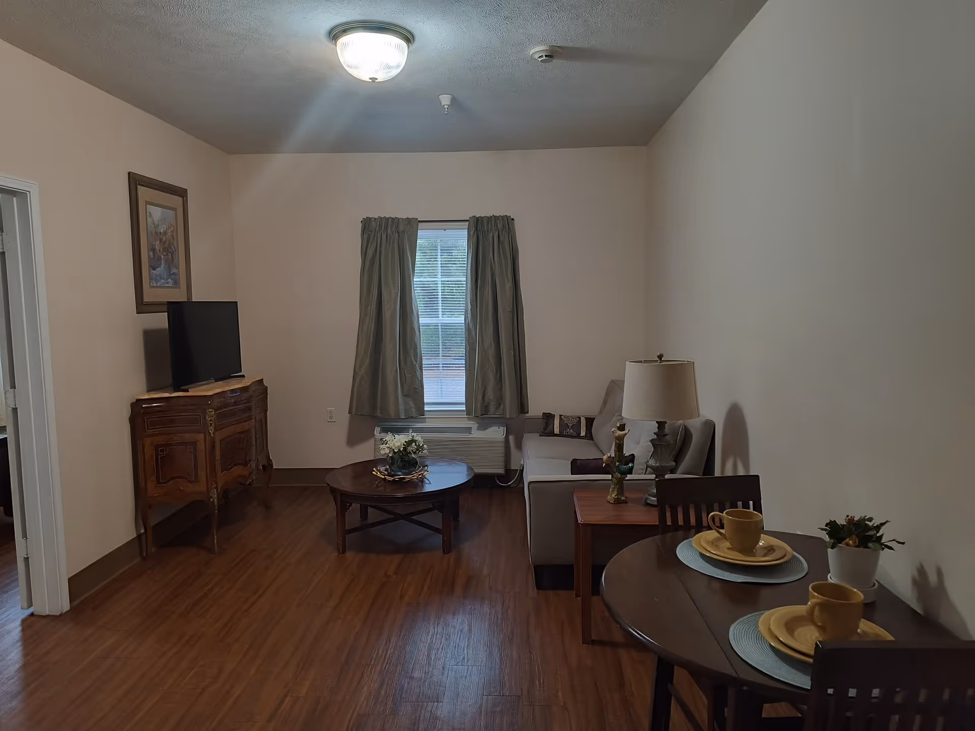 A cozy living room with wooden flooring, a small round dining table set for two with yellow cups and plates, a beige sofa with cushions, a wooden coffee table with a flower arrangement, a wooden cabinet with a flat-screen TV on top, a window with green curtains, and a ceiling light fixture.