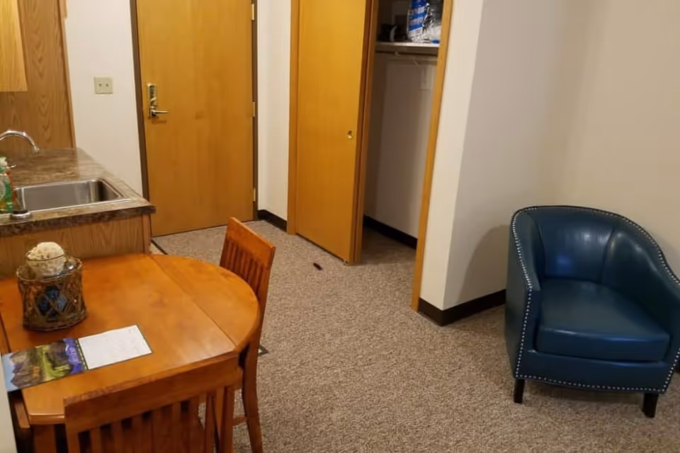 Small senior living unit interior showing a kitchenette with a sink, a round wooden dining table and chairs, an open closet and a blue armchair near the entry door.