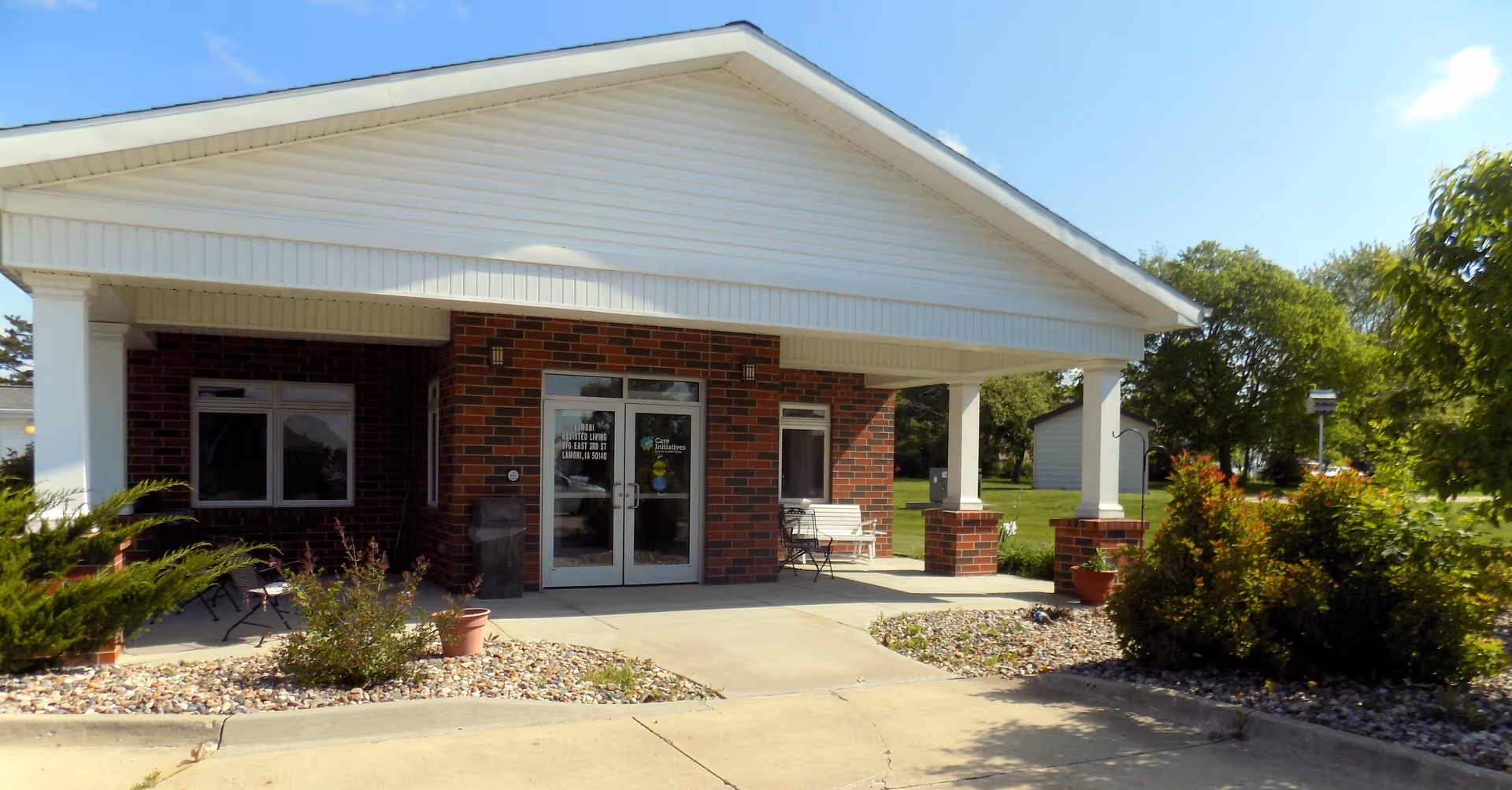 Front exterior view of a single-story brick building with a white gabled roof and a covered entrance supported by white columns. There are glass double doors at the entrance, a bench and chairs on the porch, and landscaping with bushes and rocks around the walkway. The sky is clear and blue.