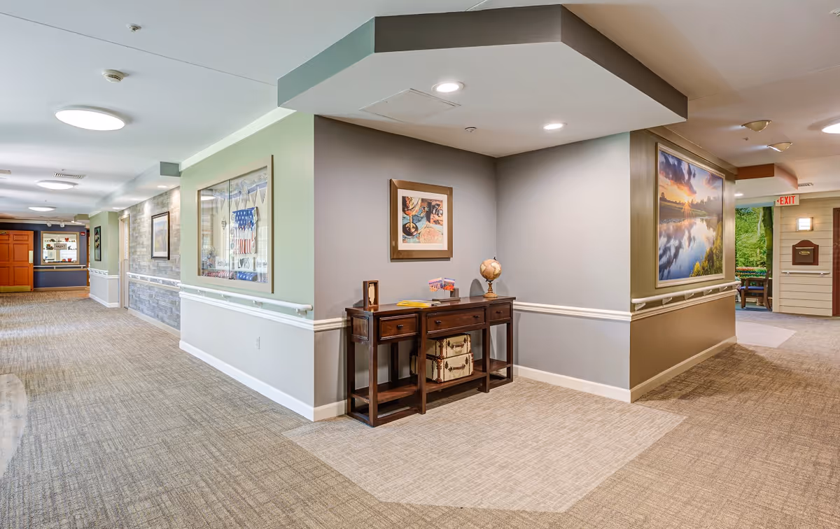 Well-lit interior hallway of a senior living facility with a wooden console table, framed artwork, and carpeted floors.
