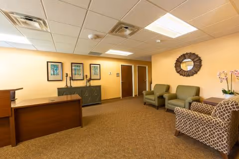 Warmly lit reception/waiting area with a wooden desk, upholstered chairs, decorative wall art, and a carpeted floor.