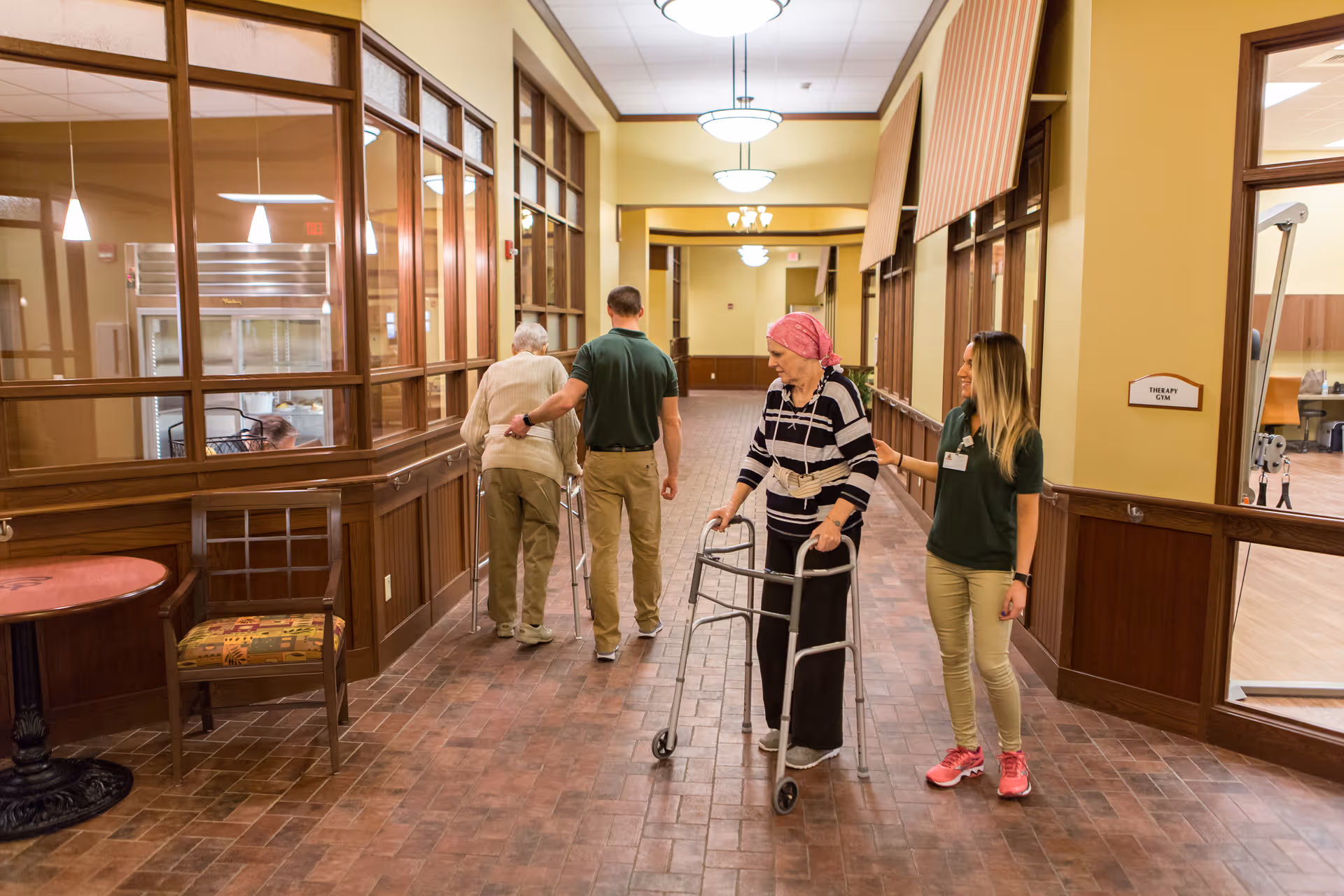 Two elderly individuals using walkers are assisted by two caregivers as they walk down a wide hallway with wooden paneling and large windows. The hallway has a warm, inviting atmosphere with tiled floors and overhead lighting. A sign on the right indicates a therapy gym nearby.