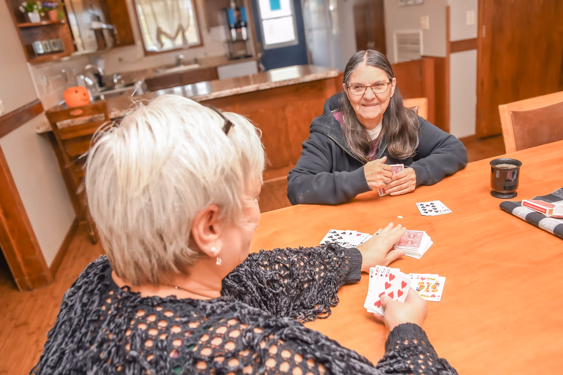 Two elderly women sitting at a wooden table playing cards in a cozy kitchen or dining area. One woman with short white hair is facing away from the camera, while the other woman with long dark hair and glasses is smiling and holding cards. The background shows kitchen cabinets, a countertop, and a window with curtains.