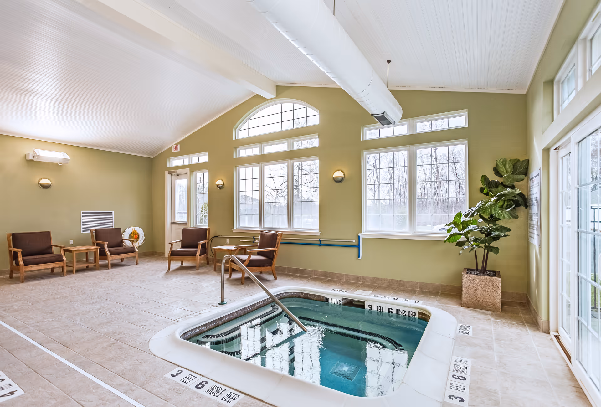 Indoor pool area with a small rectangular pool or hot tub surrounded by beige tiled floor. There are four wooden chairs with brown cushions and two small tables along the green walls. Large windows with white frames let in natural light, and a potted plant is placed near a glass door on the right side. The ceiling is white with exposed ductwork.