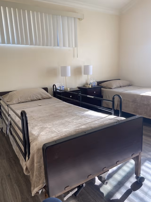 Two single hospital-style beds with side rails, nightstands and lamps between them in a bright, sparsely furnished assisted-living bedroom.