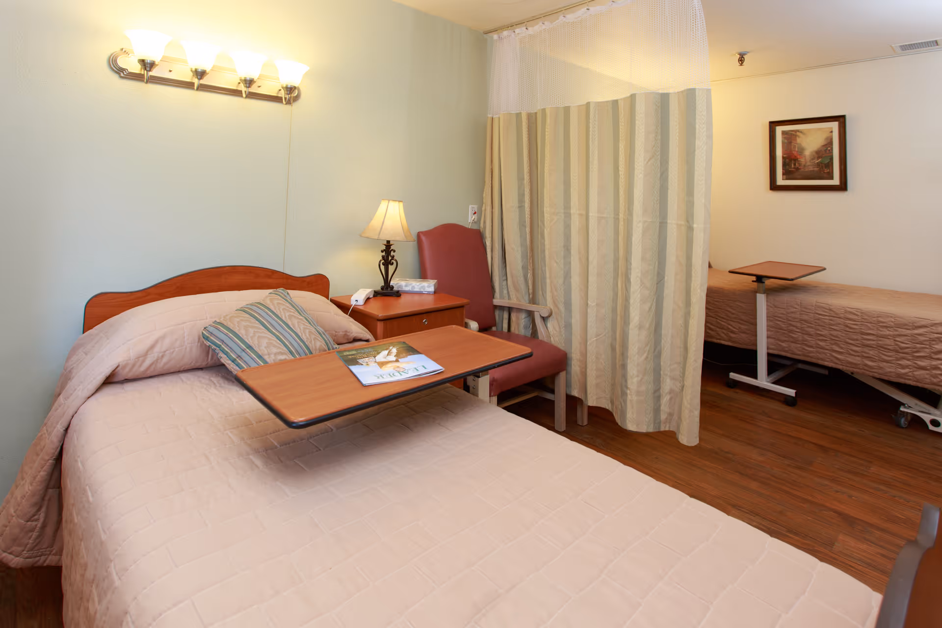 A senior care bedroom with two beds separated by a privacy curtain, overbed tables, a bedside lamp, and a chair.