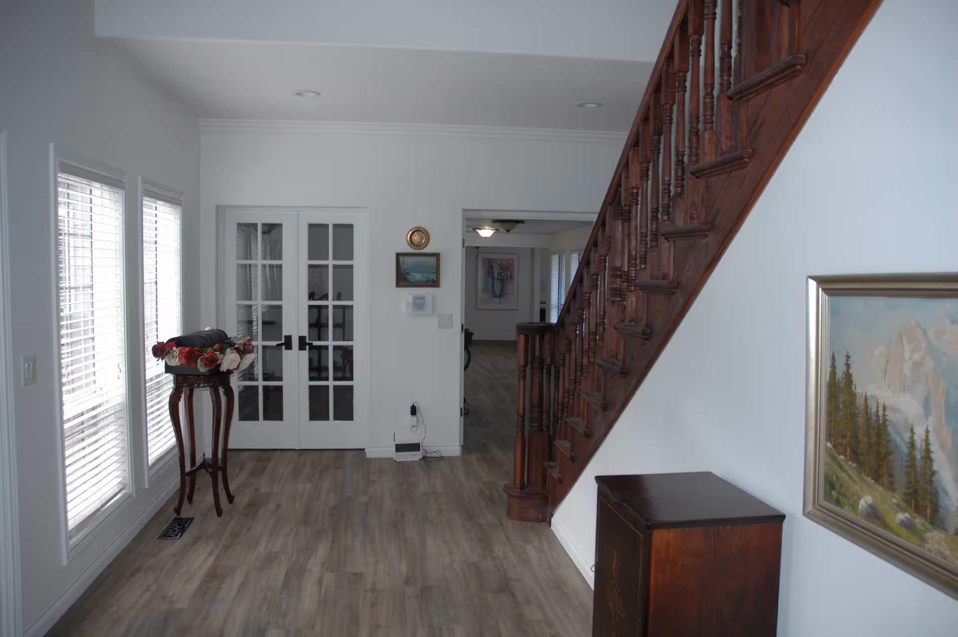 Interior view of a hallway with wooden stairs on the right, a small wooden cabinet below a framed painting of a mountain landscape, and a tall wooden stand with a floral arrangement near two large windows with blinds. Double glass doors and an open doorway lead to another room in the background.