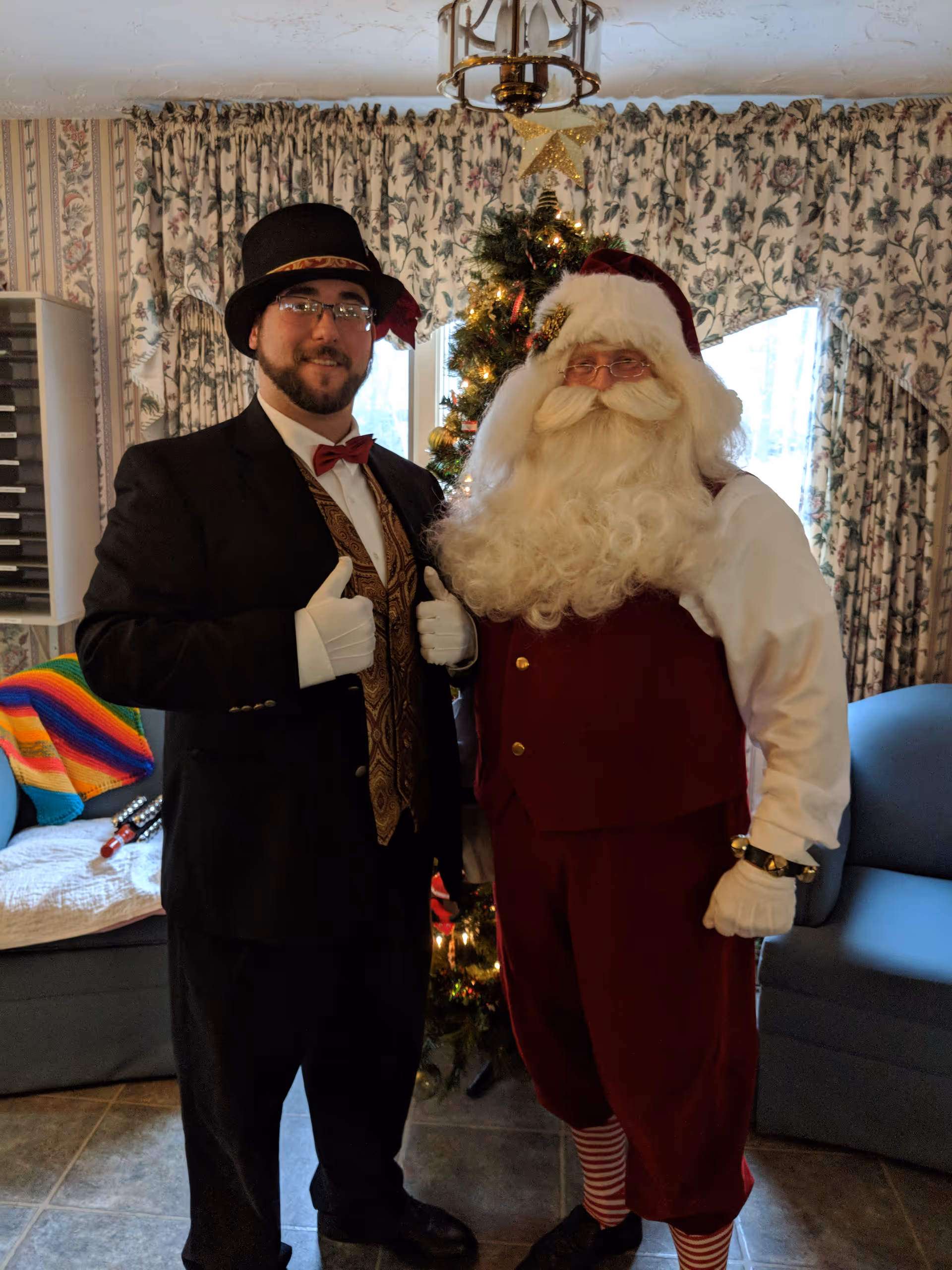 Two men standing indoors in front of a decorated Christmas tree. One man is dressed as Santa Claus with a red velvet vest, white shirt, white gloves, and a Santa hat. The other man is wearing a black suit with a patterned gold vest, red bow tie, white gloves, and a black top hat. The room has floral curtains, a blue armchair, and a colorful blanket on a couch.