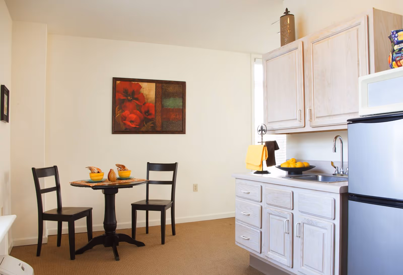 Small kitchenette and dining nook with a round table and two chairs, cabinets, sink, and a refrigerator under a framed wall painting.