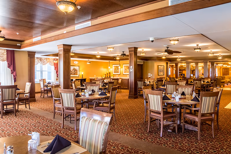 A spacious dining room in a senior living facility with multiple round wooden tables and chairs arranged neatly. Each table is set with black folded napkins, salt and pepper shakers, and small condiment containers. The room features warm yellow walls, carpeted floors with a patterned design, wooden columns, and ceiling lights. Large windows with draped curtains allow natural light to enter the space.
